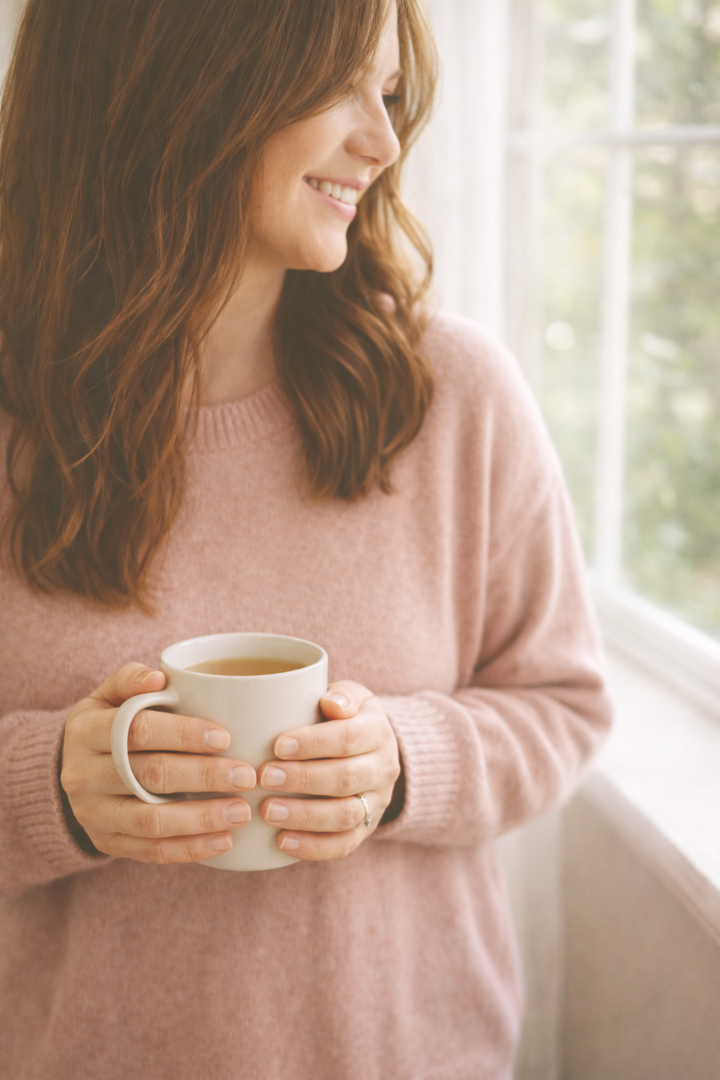 Woman with long brunette hair standing by a window holding a mug, soft natural light creating a calm and peaceful moment